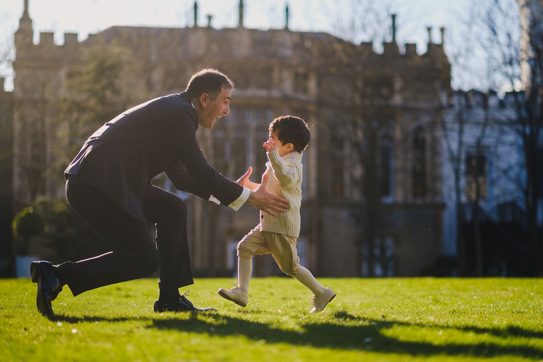 Little boy leaping into his dad's arms in the sunshine, Groom greeting guests with a hug, by Strawberry Hill House wedding photographer Owen Billcliffe